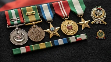 Realistic illustration of the military medals and badges awarded to Ali Al-Fadhli, including Kuwait Military Wounded Medal, National Guard Duty Medal, Kuwait Liberation Star, Kuwait Military Bravery Medal, Kuwait Liberation Medal, shown against a light Kuwait flag background. AI-generated illustration, not an authentic photograph.