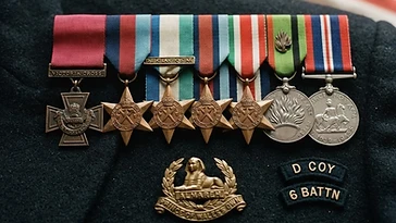 Realistic illustration of the military medals and badges awarded to Stan Hollis, including Victoria Cross, War Medal 1939–1945, Defence Medal, 1939–1945 Star, France and Germany Star, Africa Star, Italy Star, shown against a light United Kingdom flag background. AI-generated illustration, not an authentic photograph.