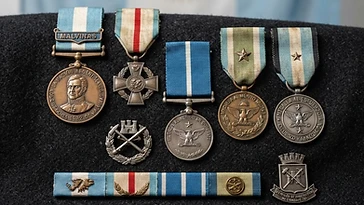 Realistic illustration of the military medals and badges awarded to Juan Jose Gomez Centurion, including Killed in Combat Medal, Medal for the Malvinas Campaign 1982, Wounded in Combat Medal, Army Military Merit Medal, Army Effort and Abnegation Medal, shown against a light Argentina flag background. AI-generated illustration, not an authentic photograph.