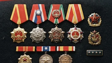 Realistic illustration of the military medals and badges awarded to Zhu De, including Order of Bayi (1955), Order of Independence and Freedom, Order of Liberation, Order of the Resplendent Banner, shown against a light China flag background. AI-generated illustration, not an authentic photograph.