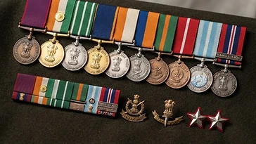 Realistic illustration of the military medals and badges awarded to Vikram Batra, including Param Vir Chakra, Maha Vir Chakra, Vir Chakra, Sena Medal, Siachen Glacier Medal, Samanya Seva Medal 1965, Param Vishisht Seva Medal, Shaurya Chakra, Kirti Chakra, Ashoka Chakra, shown against a light India flag background. AI-generated illustration, not an authentic photograph.