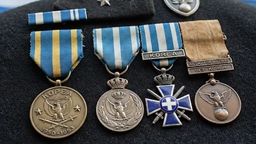 Realistic illustration of the military medals and badges awarded to Dimitrios Kopsacheilis, including Korean War Commemorative Medal, Commemorative Medal for Peacekeeping Missions, shown against a light Greece flag background. AI-generated illustration, not an authentic photograph.