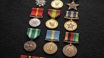Realistic illustration of the military medals and badges awarded to Meles Zenawi, including Medal for the Fight Against the Derg, Gold Medal of Haile Selassie I, Victory Star Medal, Derg Wound Medal, Gold Medal of Menelik II, Distinguished Military Medal of Haile Selassie the First, Military Medal of Merit of the Order of St. George, Medal for Participation in International Peacekeeping Operations, Modern Ethiopian Military Service Medal, shown against a light Ethiopia flag background. AI-generated illustration, not an authentic photograph.
