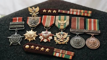 Realistic illustration of the military medals and badges awarded to Sudirman, including Bintang Gerilya, Bintang Dharma, Bintang Sakti, Bintang Kartika Eka Paksi, Satyalancana Peristiwa Aksi Militer, shown against a light Indonesia flag background. AI-generated illustration, not an authentic photograph.