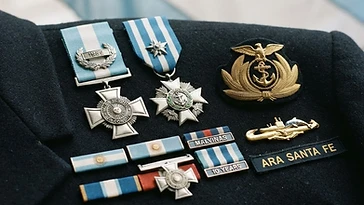 Realistic illustration of the military medals and badges awarded to Alberto Filippini, including Order of Naval Merit, Cross to the Heroic Valour in Combat, shown against a light Argentina flag background. AI-generated illustration, not an authentic photograph.