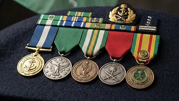 Realistic illustration of the military medals and badges awarded to Fahd bin Abdullah, including Naval Forces Medal, Medal of the Falcon of Aviation, Medal of Military Appreciation, Combat Medal, Saudi Kuwait Liberation Medal, shown against a light Saudi Arabia flag background. AI-generated illustration, not an authentic photograph.