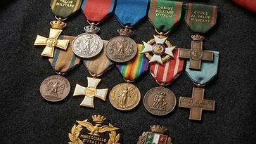 Realistic illustration of the military medals and badges awarded to Giovanni Messe, including War Cross of Military Valor, Commemorative Medal of Military Operations in East Africa, Order of Vittorio Veneto, Allied Victory Medal, Commemorative Medal for the Italo-Austrian War 1915-1918, War Merit Cross, Military Order of Italy, Bronze Medal of Military Valor, Silver Medal of Military Valor, Gold Medal of Military Valor, shown against a light Italy flag background. AI-generated illustration, not an authentic photograph.