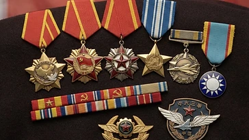 Realistic illustration of the military medals and badges awarded to Wang Hai, including Chinese People's Volunteers Resist America and Aid Korea Memorial Medal, National Defense Service Medal, Medal of Performing Combat Mission, August 1 Medal, Order of Blue Sky and White Sun, Order of National Glory, shown against a light China flag background. AI-generated illustration, not an authentic photograph.