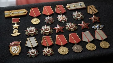 Realistic illustration of the military medals and badges awarded to Ivan Kozhedub, including Gold Star Medal, Medal for the Victory over Germany in the Great Patriotic War 1941–1945, Medal for the Defence of Stalingrad, Medal for Battle Merit, Order of Glory, Order of the Patriotic War, Order of the Red Star, Order of the Red Banner, shown against a light Soviet Union flag background. AI-generated illustration, not an authentic photograph.