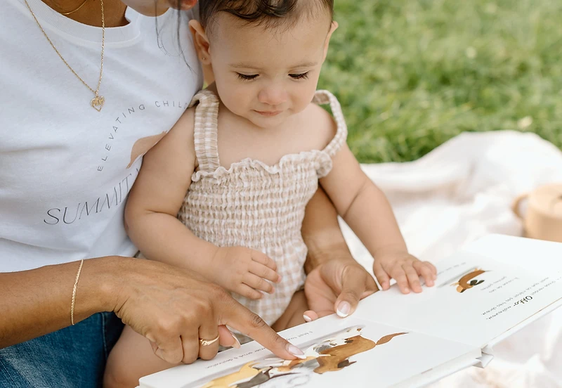 A nanny and child reading together near a sunlit window with mountains in the distance