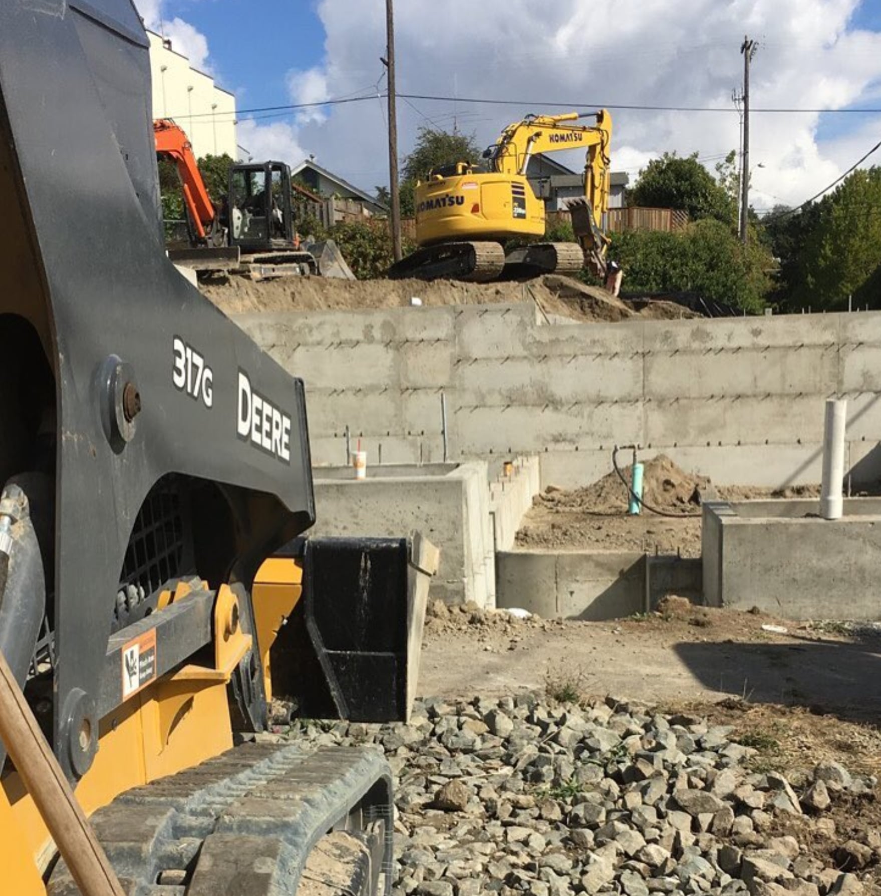 Deere skid steer on gravel with a Komatsu excavator behind a retaining wall