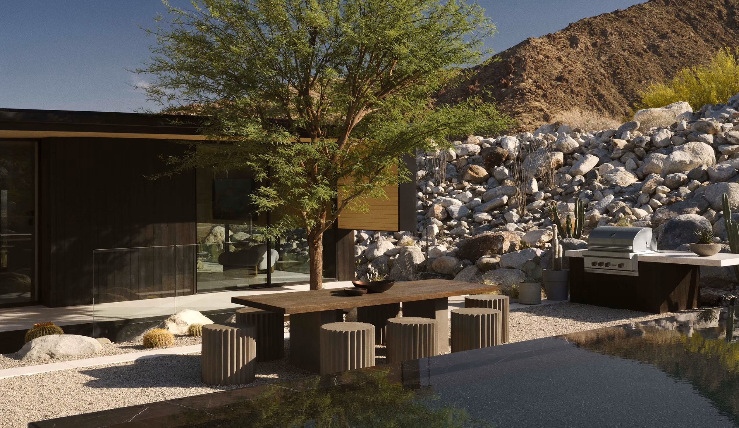 Outdoor courtyard dining area with a long wooden table, stools, and an outdoor kitchen against a boulder-strewn slope.