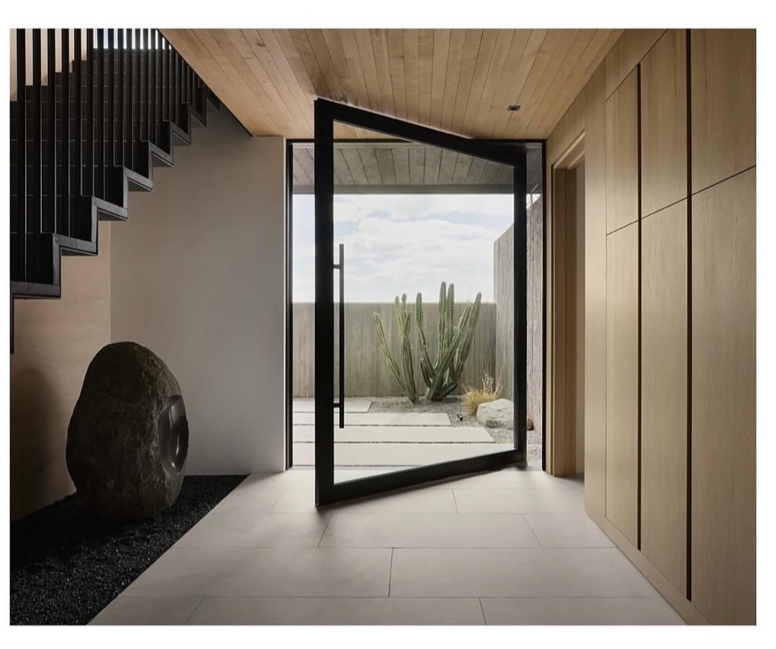 Entry-hall pivot door opening to a desert garden with concrete stepping stones and a cactus.