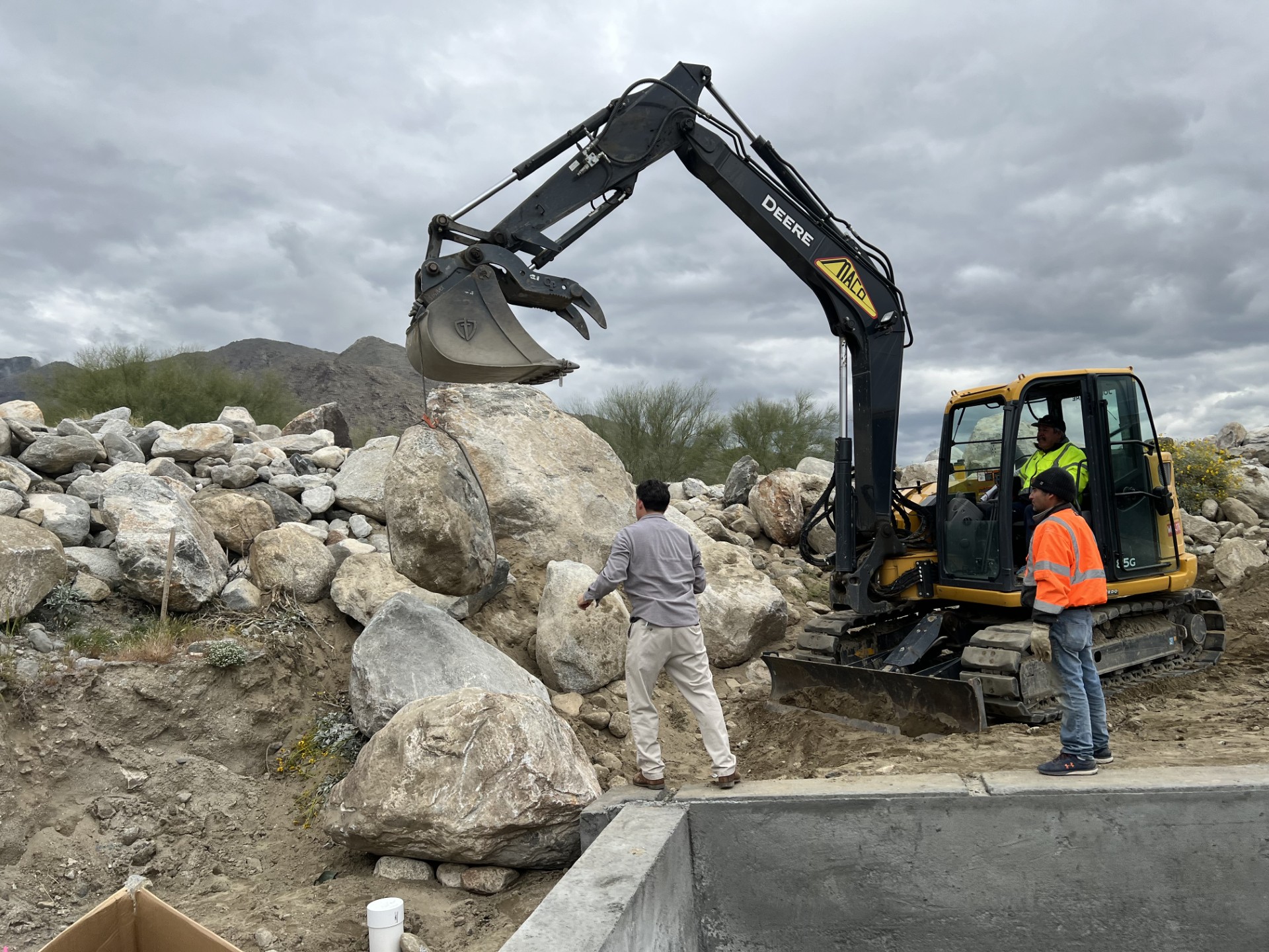 Excavator lifting a large boulder while a team member directs its placement on site.