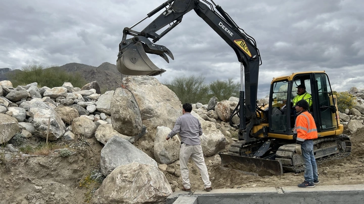 Excavator lifting a large boulder while a team member directs its placement on site