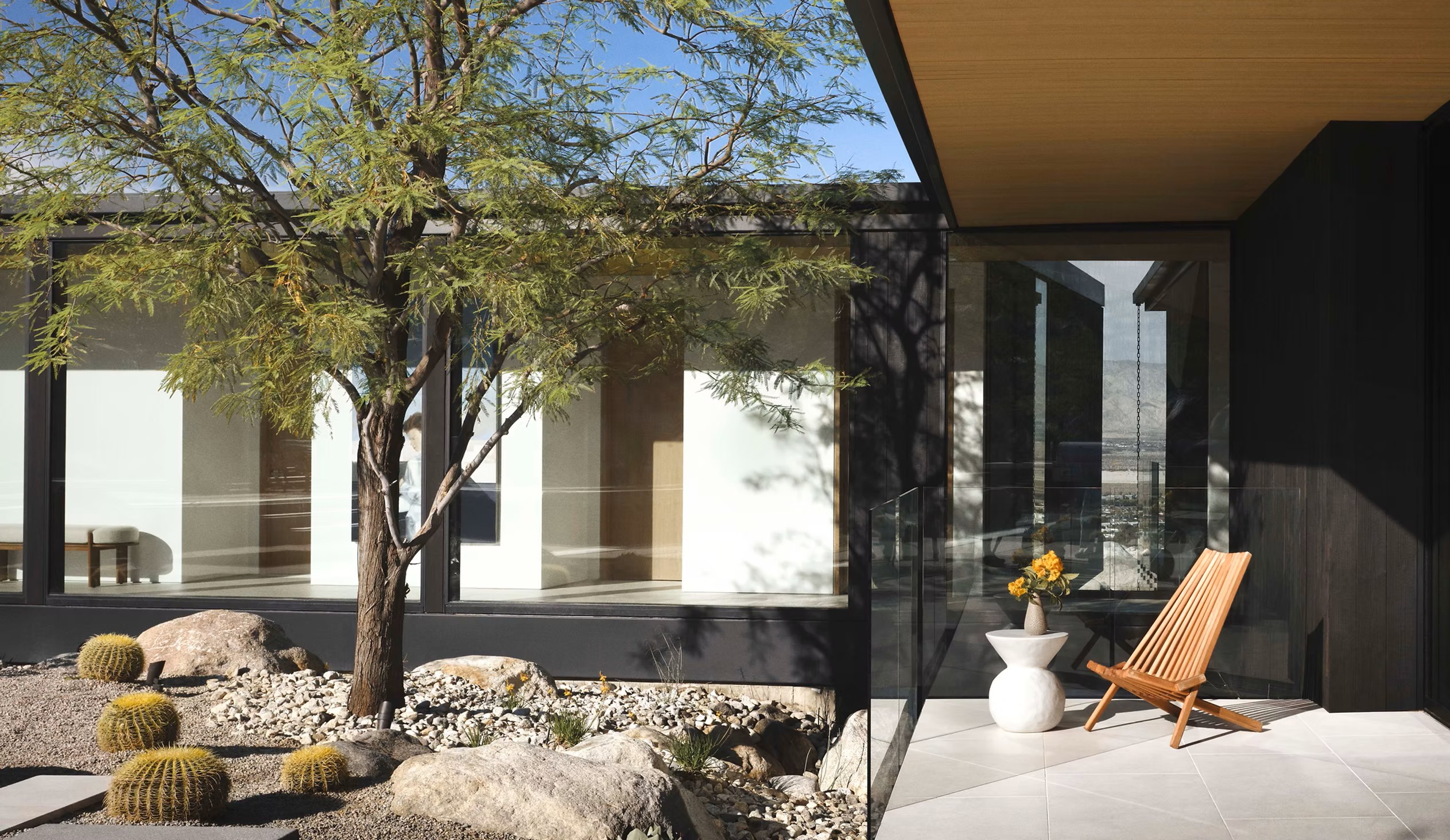 Shaded courtyard with a mature tree, golden barrel cacti, and a lounge chair beside dark wood-clad architecture.