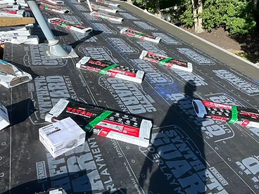TAMKO Synthetic Guard underlayment with staged bundles of TITAN XT premium architectural shingles and a metal chimney pipe boot on a roof deck
