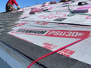 Roofer in a red shirt kneeling on TITANIUM PSU30 underlayment using a pneumatic nail gun with a black starter strip along the eave and a green air hose running across the roof