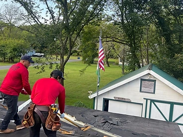 Two workers in red shirts using long-handled shingle removers to strip old asphalt shingles from a residential roof