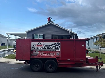 Red Veterans Roofing debris trailer parked on the street in front of a grey house with workers on the roof