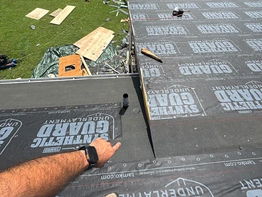 Worker's hand pointing toward an exposed edge of TAMKO Synthetic Guard black underlayment near a small pipe penetration on a roof deck