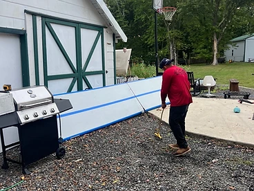 Roofer in a red shirt operating a rolling blue magnetic sweeper over gravel alongside a white-sided home with a green-trimmed garage door