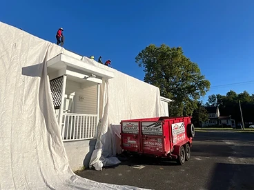 Red Veterans Roofing debris trailer positioned next to a white house with a protective tarp draped down the siding and workers on the roof