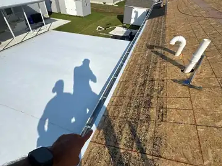 First-person view from the roof pointing at a bright metal drip edge installed along the eave over exposed OSB decking with two PVC vent pipes