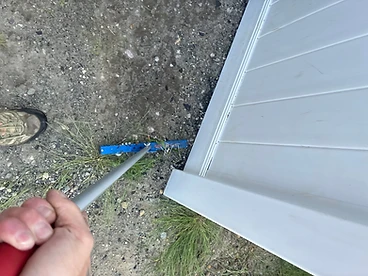 Blue magnetic sweeper head with collected roofing nails pressed flat against dirt and gravel alongside a white-sided home foundation wall