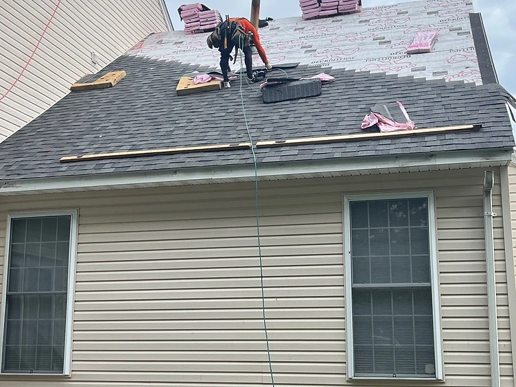 Worker in an orange shirt and safety harness kneeling on a roof nailing architectural shingles over Deck Defense underlayment with the lower section already shingled