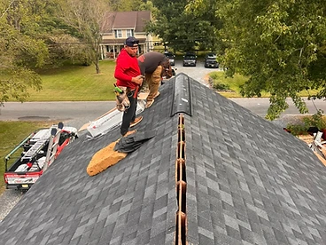 Two roofers installing a black ridge vent on the peak of a grey shingled roof with a ladder visible and a suburban background