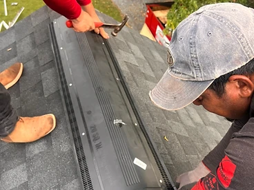 Two roofers installing a black plastic ridge vent along the peak of a grey shingled roof, one nailing with a hammer and the other positioning the vent