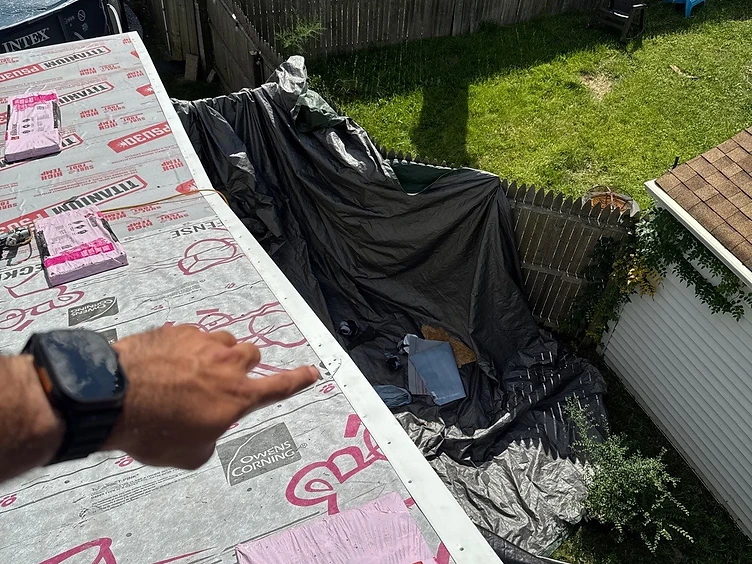 Hand pointing at a white metal rake edge drip edge installed along the sloped gable edge of a roof covered in Owens Corning Deck Defense underlayment with shingle bundles staged