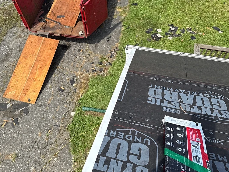 Hand pointing at a white metal rake drip edge over black TAMKO Synthetic Guard underlayment with a bundle of TAMKO TITAN XT shingles staged on the roof