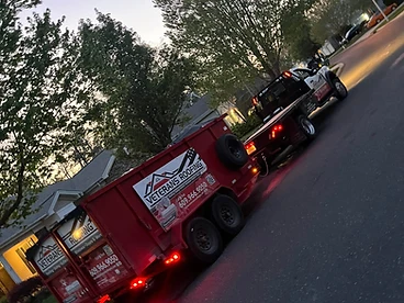 Red Veterans Roofing debris trailer being towed at dusk by a white truck with taillights illuminated
