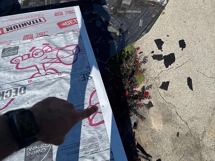Hand pointing at a white metal drip edge running along the rake of a roof covered in Owens Corning Deck Defense underlayment with a black tarp below