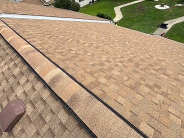 High-angle view down a completed hip and ridge cap installation on a brown architectural shingle roof with a black ridge vent visible beneath the overlapping cap shingles