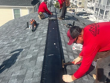 Three roofers in red Veterans Roofing shirts installing a long black ridge vent along the peak of a grey shingled roof in a suburban neighborhood