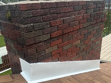 Close-up of a white metal chimney base flashing sealed with a bead of white caulk along the top edge where it meets a brick chimney above brown architectural shingles