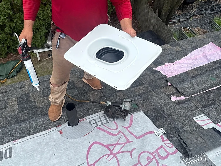 Roofer in a red shirt holding a white pipe boot flashing with sealant applied to the underside, preparing to install it over a black plumbing vent pipe on a shingled roof