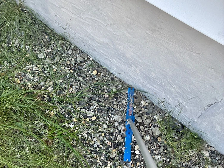 Close-up of a blue magnetic sweeper bar coated in collected rusted roofing nails being run along the base of a home's stucco foundation wall