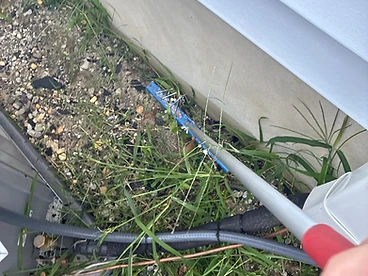Blue magnetic sweeper with a red grip handle and collected nails being run along a patch of dirt and grass beside a home's white siding and an HVAC unit
