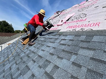 Roofer in a red shirt and straw hat using a pneumatic nail gun to install architectural shingles over white ProArmor underlayment on a clear day