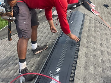 Roofer in a red shirt kneeling on a grey shingled roof installing a black CertainTeed ridge vent along the peak with a red cord running across the roof
