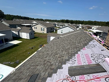 High-angle view down a roof slope partially covered in multi-tonal grey architectural shingles with ProArmor underlayment still exposed above and shingle bundles staged