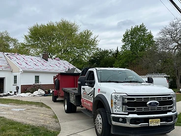 White Ford Super Duty truck hitched to a red debris trailer parked on a driveway beside a house mid-roof with pink underlayment visible