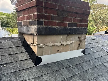 Brick and cinder block chimney on a charcoal shingled roof with black metal step flashing along the sides and white metal apron flashing sealed against the front face