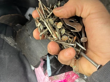 Close-up of a hand holding a large collection of rusted roofing nails and a metal scraper tool inside a black trash bag during cleanup