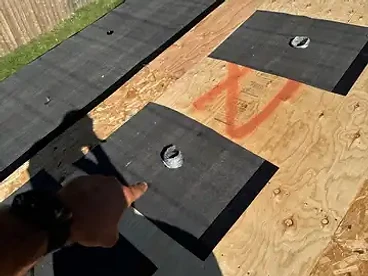 Two black underlayment patches surrounding circular pipe vents on OSB decking with an orange spray paint X marked between them