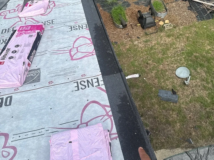 Hand pointing at a black granulated starter strip installed along the eave edge of a roof over Owens Corning Deck Defense underlayment with a worker installing shingles above